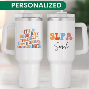 May include: Two white personalized travel mugs with lids and handles. The mug on the left has the text "It's a good day to help tiny humans communicate". The mug on the right has the text "SLPA Sarah".