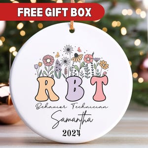 May include: White ceramic ornament with a floral design and the letters RBT, standing for Behavior Technician. The ornament also includes the name Samantha and the year 2024.