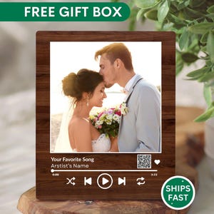 May include: A wooden plaque featuring a wedding photo of a couple, designed to resemble a music player interface. The plaque includes the text "Your Favorite Song" and "Artist's Name", along with a play button and a QR code. The words "FREE GIFT BOX" are at the top.