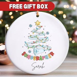 May include: White ceramic Christmas ornament with a colorful cartoon illustration of a Christmas tree with lights and a sonogram probe. The text "Sonographer" is written in rainbow letters and the name "Sarah" is written below.