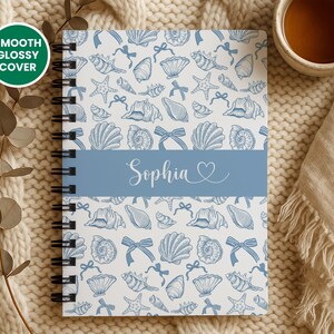 K&ouml;nnte beinhalten: Ein spiralgebundenes Notizbuch mit wei&szlig;em Hintergrund und einem Muster aus blauen Muscheln, Schleifen und Seesternen. Ein blaues Band in der Mitte zeigt den Namen "Sophia" in Schreibschrift. Das Cover ist mit "Smooth Glossy Cover" beschriftet.