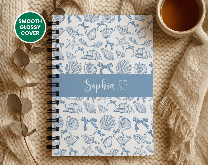 K&ouml;nnte beinhalten: Ein spiralgebundenes Notizbuch mit wei&szlig;em Hintergrund und einem Muster aus blauen Muscheln, Schleifen und Seesternen. Ein blaues Band in der Mitte zeigt den Namen "Sophia" in Schreibschrift. Das Cover ist mit "Smooth Glossy Cover" beschriftet.