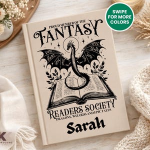 May include: A beige book cover with a black dragon illustration and the text "PROUD MEMBER OF THE FANTASY READERS SOCIETY". The book also has the name "Sarah" printed on it. The book is a fantasy-themed item.