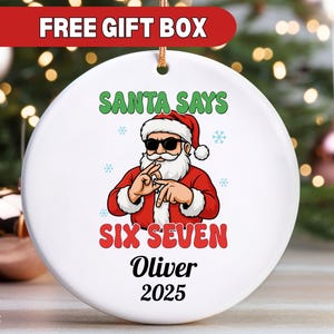Könnte beinhalten: Weißer, runder Weihnachtsschmuck mit einem Santa Claus-Motiv mit Sonnenbrille. Der Text "SANTA SAYS SIX SEVEN" steht über "Oliver 2025". Der Anhänger hat eine goldene Schnur zum Aufhängen.