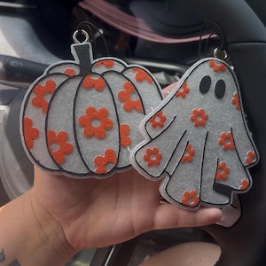 May include: Two Halloween-themed car air fresheners. One is a pumpkin shape, the other a ghost. Both are gray with orange flower patterns and black outlines. They have black loops for hanging.