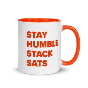 Puede incluir: Taza de cerámica blanca con borde y asa naranja. La taza tiene el texto "STAY HUMBLE STACK SATS" impreso en naranja.