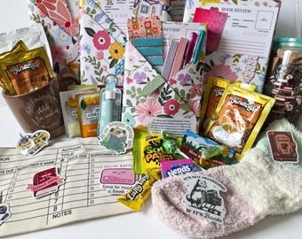 May include: A gift set featuring floral-patterned book review journals, a brown mug with the text 'shhh, I'm reading', various coffee packets, colourful pens, stickers, sweets, a library checkout card pouch, and a pair of soft, multi-coloured fuzzy socks.