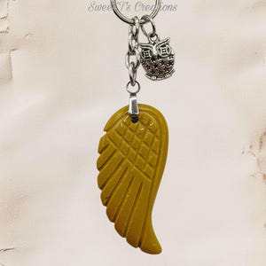 May include: A keychain featuring a mustard yellow wing-shaped pendant with a grid-like texture on the upper portion. Attached to the silver-toned metal ring is a small silver owl charm with star details. The text Sweet T's Creations is visible.