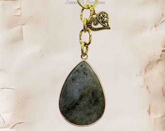 May include: A teardrop-shaped pendant featuring a grey, mottled stone set in a gold-toned metal frame. The pendant hangs from a gold-toned chain with a small, ornate heart-shaped charm attached near the top. The text Sweet T's Creations is visible.
