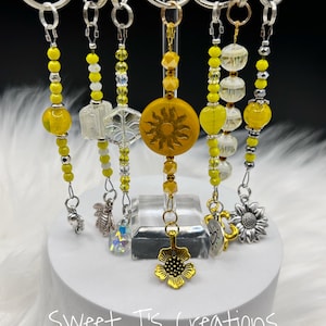May include: Six yellow and silver beaded keychains with different charms. The charms include a bee, a sunflower, a sun, a heart, a crystal, and a snowflake. The keychains are displayed on a white stand.
