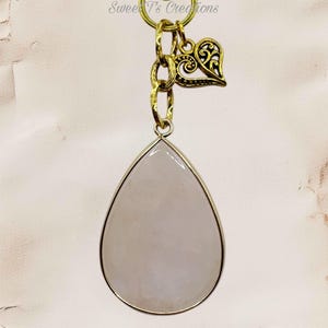 May include: A teardrop-shaped pendant featuring a smooth, pale pink stone set in a gold-toned metal frame. The pendant hangs from a gold-toned chain link with an attached filigree heart charm. The text Sweet T's Creations is visible at the top.