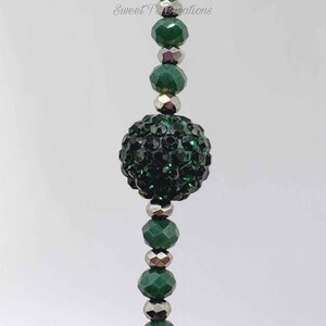 May include: A vertical strand of beads featuring alternating dark green faceted crystal beads and smaller metallic silver-toned spacer beads, centered by a large, round, dark green rhinestone-encrusted bead. The text Sweet T Creations is visible.
