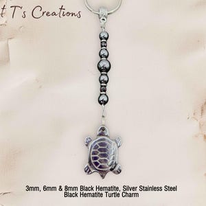 May include: A turtle charm keychain. The charm is made of black hematite and silver stainless steel. The keychain also includes black hematite beads. The text "Sweet T's Creations" is at the top.