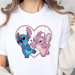 Puede incluir: Camiseta blanca con un gr&aacute;fico de Stitch y Angel de Lilo & Stitch. Los personajes est&aacute;n dentro de un coraz&oacute;n rojo con un patr&oacute;n de cuadr&iacute;cula.