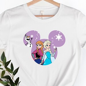 May include: White t-shirt with a purple Mickey Mouse silhouette featuring Anna and Elsa from Frozen and Olaf. The characters are surrounded by white snowflakes.