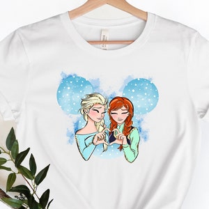 May include: White t-shirt with a graphic of Elsa and Anna from Frozen making a heart shape with their hands. The graphic is surrounded by a blue watercolor background with white snowflakes.