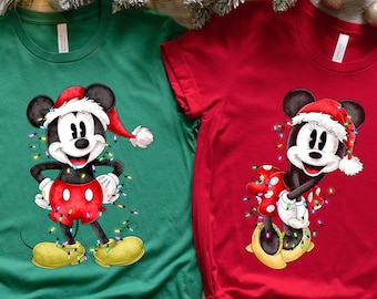 May include: Two Christmas themed t-shirts featuring Mickey Mouse and Minnie Mouse wearing Santa hats and surrounded by colorful Christmas lights. The Mickey Mouse shirt is green and the Minnie Mouse shirt is red.