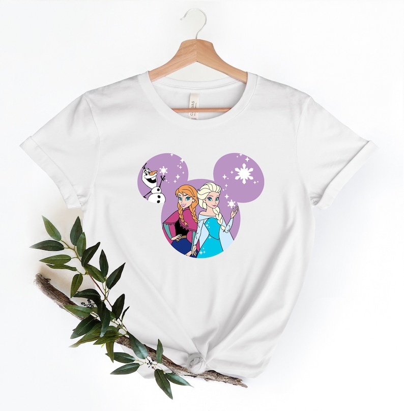 May include: White t-shirt with a purple Mickey Mouse silhouette featuring Anna and Elsa from Frozen and Olaf. The characters are surrounded by white snowflakes.