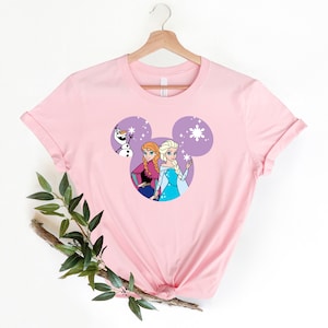 May include: Pink t-shirt with a purple Mickey Mouse head design featuring Anna and Elsa from Frozen and Olaf. The design includes snowflakes.