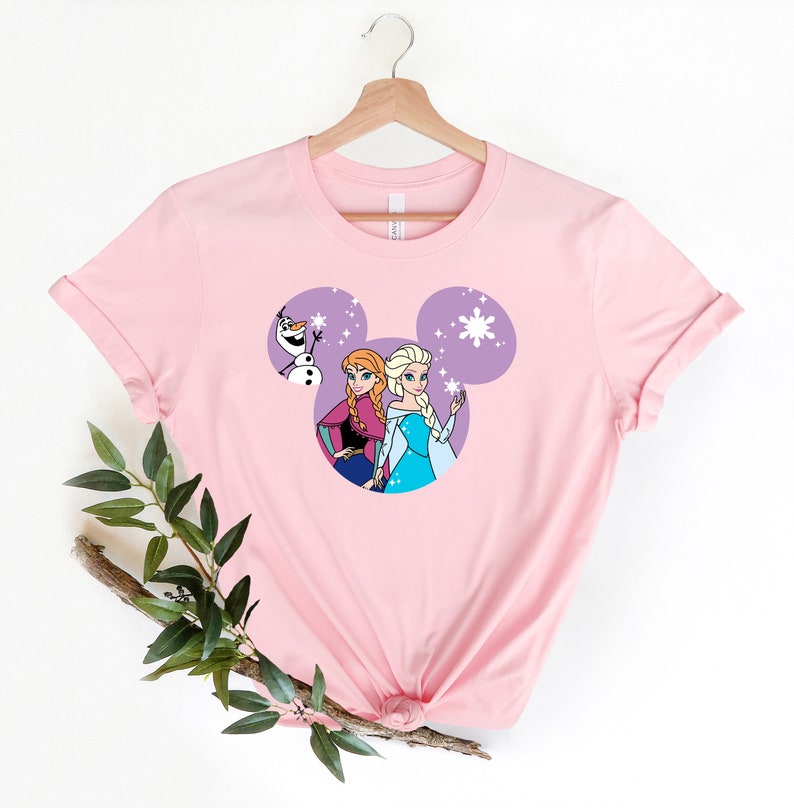May include: Pink t-shirt with a purple Mickey Mouse head design featuring Anna and Elsa from Frozen and Olaf. The design includes snowflakes.