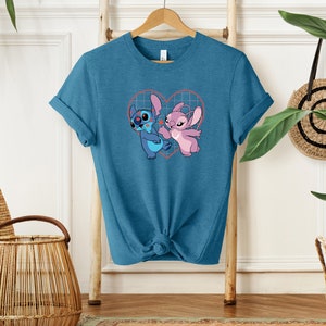 Puede incluir: Una camiseta azul turquesa con un gr&aacute;fico de Stitch y Angel de la pel&iacute;cula de Disney Lilo & Stitch. El gr&aacute;fico muestra a Stitch y Angel dentro de un coraz&oacute;n rosa con un patr&oacute;n de cuadr&iacute;cula.