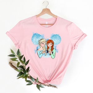 May include: Pink t-shirt with a graphic of Elsa and Anna from Frozen making a heart shape with their hands. The graphic is set against a blue and white watercolor background with snowflakes.