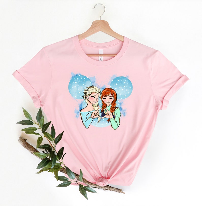 May include: Pink t-shirt with a graphic of Elsa and Anna from Frozen making a heart shape with their hands. The graphic is set against a blue and white watercolor background with snowflakes.