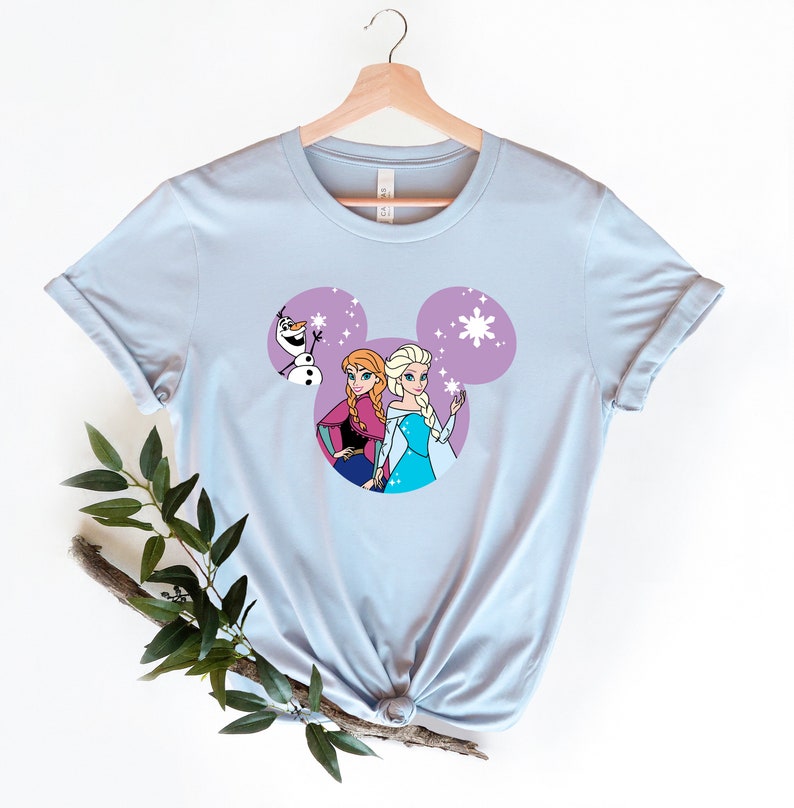 May include: Light blue t-shirt with a purple Mickey Mouse head design featuring the characters Elsa and Anna from Frozen and Olaf.