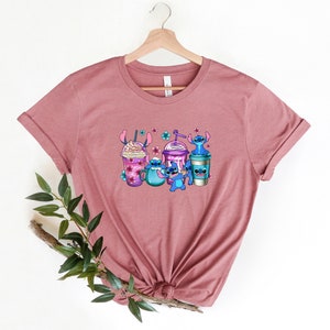 May include: A mauve t-shirt with a graphic print of Stitch from Lilo & Stitch holding different types of coffee drinks. The graphic features Stitch in blue, purple, and pink colors.