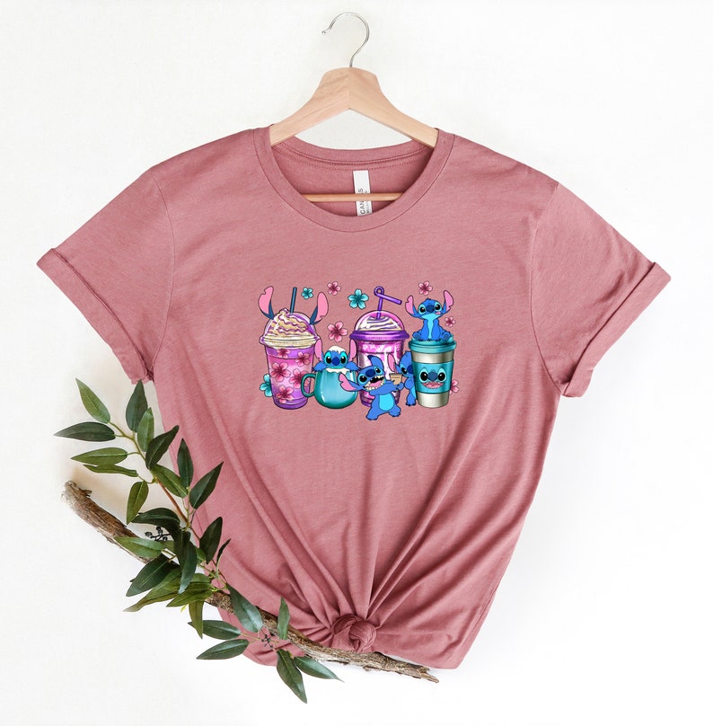 May include: A mauve t-shirt with a graphic print of Stitch from Lilo & Stitch holding different types of coffee drinks. The graphic features Stitch in blue, purple, and pink colors.