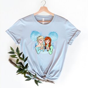 May include: Light blue t-shirt with a graphic of the Disney characters Elsa and Anna from Frozen, making a heart shape with their hands. The graphic is set against a blue and white watercolor background with snowflakes.