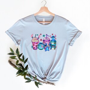 May include: Light blue t-shirt with a graphic of Stitch characters from the Disney movie Lilo & Stitch. The characters are holding various colorful drinks with pink flowers and a blue background.