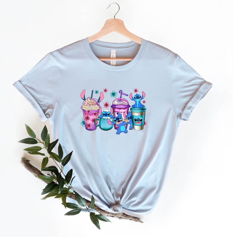 May include: Light blue t-shirt with a graphic of Stitch characters from the Disney movie Lilo & Stitch. The characters are holding various colorful drinks with pink flowers and a blue background.