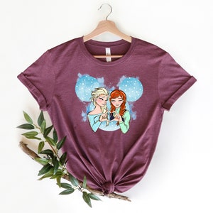 May include: A maroon t-shirt with a graphic of Elsa and Anna from Frozen, making a heart shape with their hands. The graphic is set against a blue and white watercolor background with a Mickey Mouse ear silhouette.