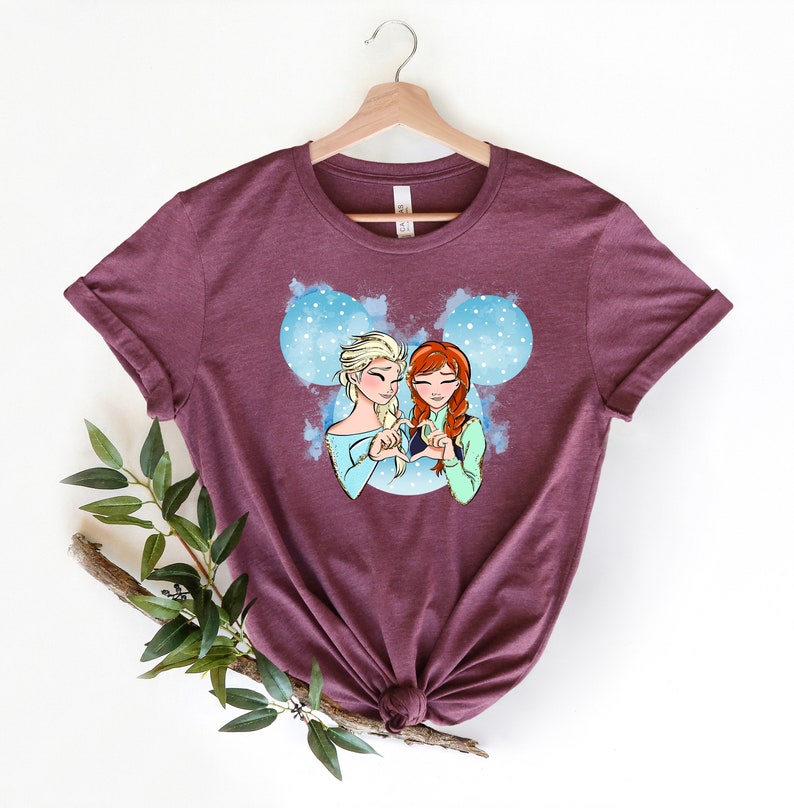 May include: A maroon t-shirt with a graphic of Elsa and Anna from Frozen, making a heart shape with their hands. The graphic is set against a blue and white watercolor background with a Mickey Mouse ear silhouette.
