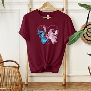 Puede incluir: Una camiseta de color granate con un gr&aacute;fico de Stitch y Angel en una cuadr&iacute;cula en forma de coraz&oacute;n. Los personajes son azules y rosas, y el coraz&oacute;n es rojo.