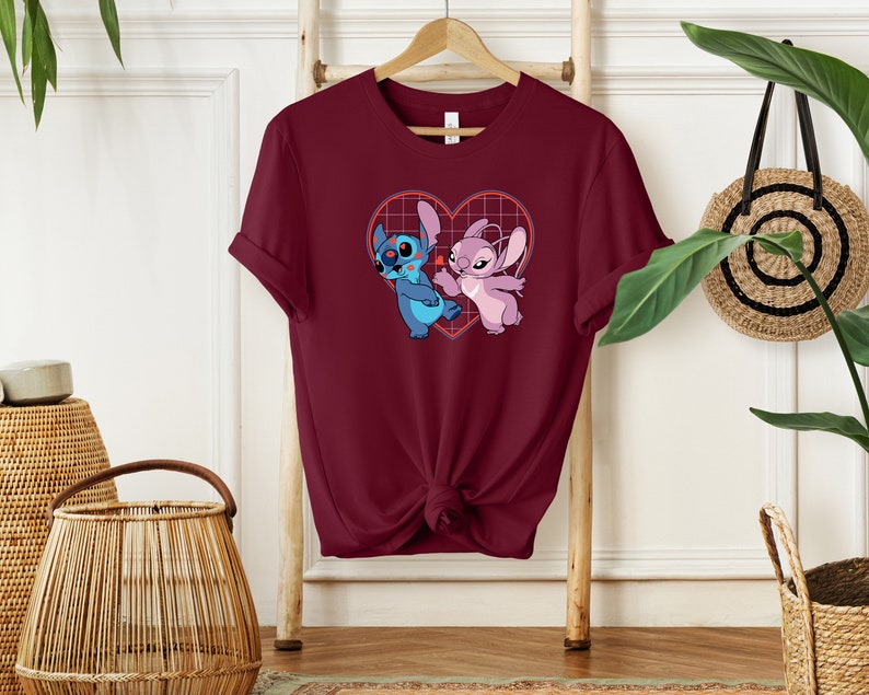 Puede incluir: Una camiseta de color granate con un gr&aacute;fico de Stitch y Angel en una cuadr&iacute;cula en forma de coraz&oacute;n. Los personajes son azules y rosas, y el coraz&oacute;n es rojo.