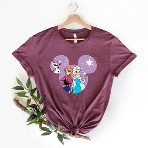 May include: A maroon t-shirt with a purple Mickey Mouse head design featuring Elsa and Anna from Frozen and Olaf. The design includes snowflakes.