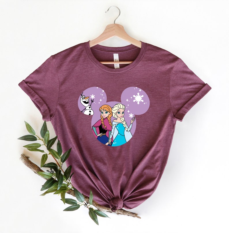 May include: A maroon t-shirt with a purple Mickey Mouse head design featuring Elsa and Anna from Frozen and Olaf. The design includes snowflakes.
