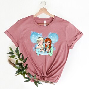 May include: A mauve t-shirt with a graphic of two cartoon girls with blonde and red hair, holding hands in the shape of a heart. The graphic is inside a blue Mickey Mouse silhouette.