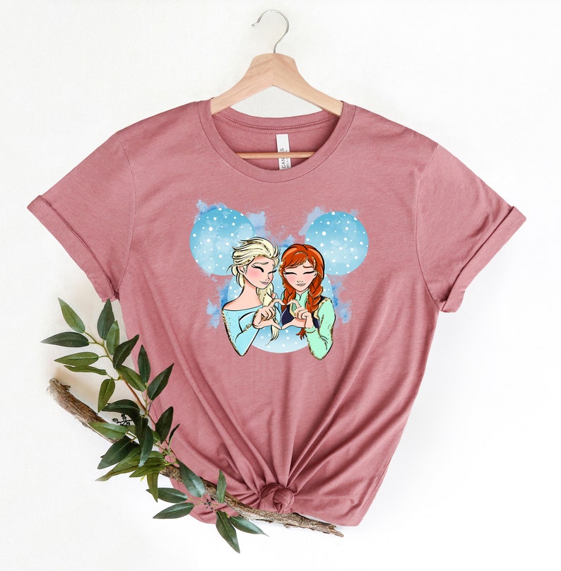 May include: A mauve t-shirt with a graphic of two cartoon girls with blonde and red hair, holding hands in the shape of a heart. The graphic is inside a blue Mickey Mouse silhouette.