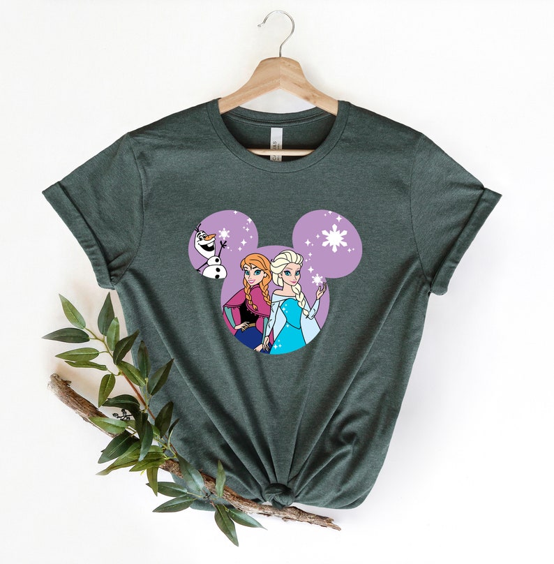 May include: A green t-shirt with a purple Mickey Mouse head design featuring Elsa and Anna from Frozen. Olaf is peeking out from behind them.