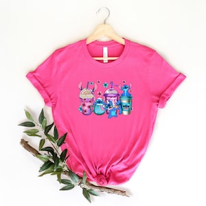 May include: Pink t-shirt with a graphic of Stitch characters holding different types of coffee drinks. The characters are blue with purple accents and are surrounded by pink flowers.