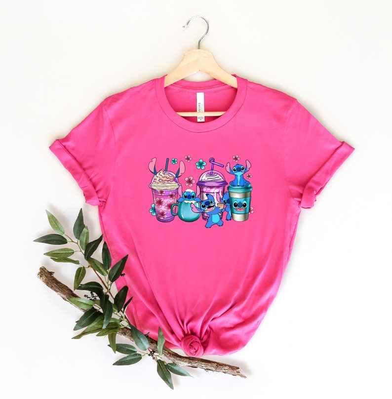May include: Pink t-shirt with a graphic of Stitch characters holding different types of coffee drinks. The characters are blue with purple accents and are surrounded by pink flowers.