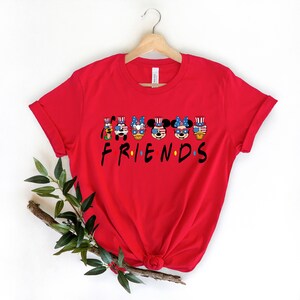 May include: Red t-shirt with a graphic of Mickey Mouse and friends wearing patriotic hats and sunglasses. The text "FRIENDS" is below the graphic.