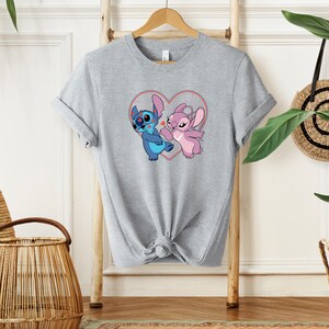 Puede incluir: Una camiseta gris con un gr&aacute;fico de Stitch y Angel de Lilo & Stitch. Los personajes est&aacute;n dentro de una forma de coraz&oacute;n con contornos rojos.