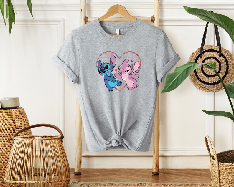 Puede incluir: Una camiseta gris con un gr&aacute;fico de Stitch y Angel de Lilo & Stitch. Los personajes est&aacute;n dentro de una forma de coraz&oacute;n con contornos rojos.