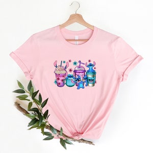 May include: Pink t-shirt with a graphic of Stitch characters holding various colorful iced coffee drinks with pink flowers.