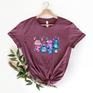 May include: A maroon t-shirt with a graphic of Stitch from Lilo & Stitch holding different coffee drinks. The graphic includes a mocha, a smoothie, and a coffee cup.