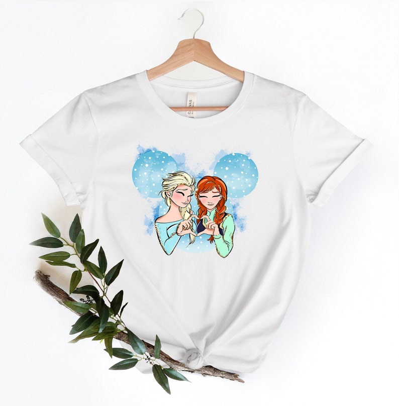 May include: White t-shirt with a graphic of Elsa and Anna from Frozen making a heart shape with their hands. The graphic is surrounded by a blue watercolor background with white snowflakes.